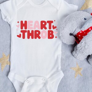 Op de afbeelding: Een witte baby romper met de tekst "HEART THROB" in roze en rode letters met hartjes rond de tekst. Een grijs olifant knuffeldier ligt op de romper.