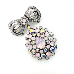 May include: A silver-tone brooch featuring a large pink teardrop crystal surrounded by smaller pink and iridescent crystals. The brooch also includes a smaller bow with iridescent crystals.