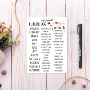 May include: A sticker sheet with a "Future Log" design for a journal, featuring monthly calendars and holiday icons. A black pen rests beside the sticker sheet on a light wood surface. Decorative greenery and twine are also present.
