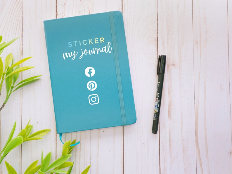 May include: A teal blue journal with the text "STICKER my journal" and social media icons for Facebook, Pinterest, and Instagram. A black pen is on a white wooden surface.