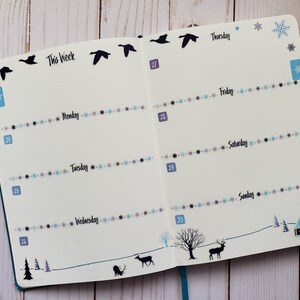 May include: A weekly planner with a winter theme. The planner has a blue cover and features a black silhouette of a flock of geese flying across the top of the page. The days of the week are listed on the right side of the page, with the dates listed on the left side. The planner is decorated with a wintery design, including snowflakes, a small house, and a deer.
