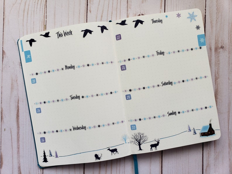 May include: A weekly planner with a winter theme. The planner has a blue cover and features a black silhouette of a flock of geese flying across the top of the page. The days of the week are listed on the right side of the page, with the dates listed on the left side. The planner is decorated with a wintery design, including snowflakes, a small house, and a deer.