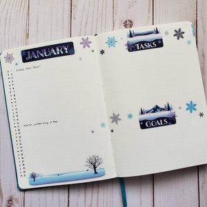 May include: An open planner with a blue cover. The left page has the word "JANUARY" written in a decorative font and a list of numbers from 1 to 31. The right page has the words "TASKS" and "GOALS" written in decorative fonts. The pages are decorated with snowflake stickers.