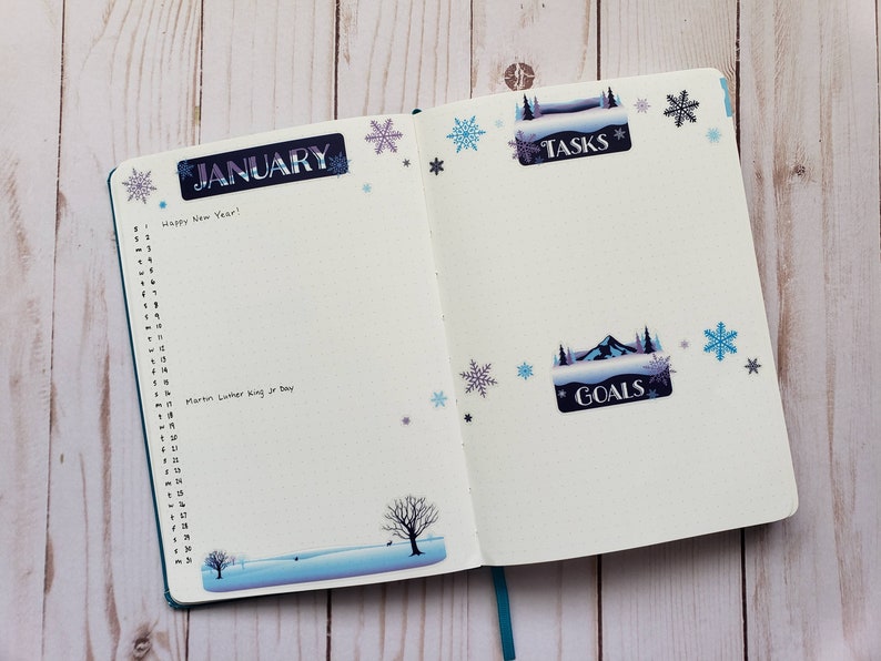 May include: An open planner with a blue cover. The left page has the word "JANUARY" written in a decorative font and a list of numbers from 1 to 31. The right page has the words "TASKS" and "GOALS" written in decorative fonts. The pages are decorated with snowflake stickers.
