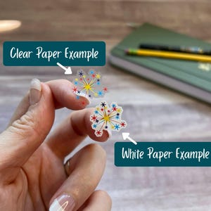 Puede incluir: Dos pegatinas peque&ntilde;as con tem&aacute;tica de fuegos artificiales y dise&ntilde;os de estrellas coloridas en rojo, azul y amarillo. Una pegatina est&aacute; impresa en papel transparente y la otra en papel blanco. Los textos indican Clear Paper Example y White Paper Example.