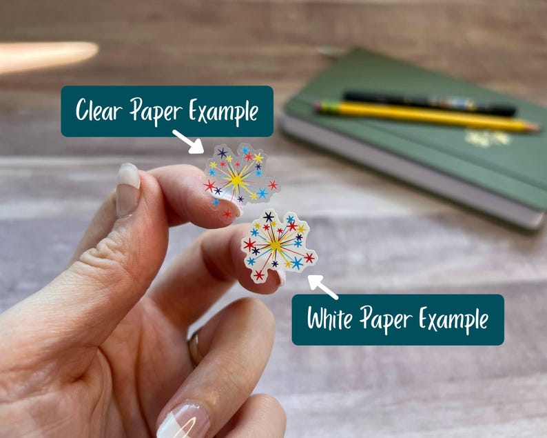 Puede incluir: Dos pegatinas peque&ntilde;as con tem&aacute;tica de fuegos artificiales y dise&ntilde;os de estrellas coloridas en rojo, azul y amarillo. Una pegatina est&aacute; impresa en papel transparente y la otra en papel blanco. Los textos indican Clear Paper Example y White Paper Example.