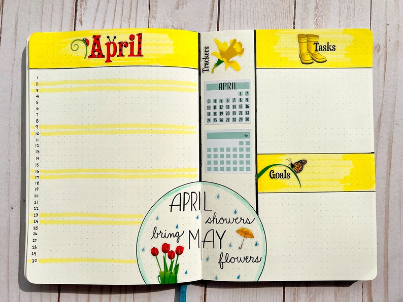 May include: A yellow and white planner spread with the word "April" in red lettering. The left page has a calendar for April and a circle with the text "April showers bring May flowers". The right page has sections for "Trackers", "Tasks", "Goals", and a blank space for writing.
