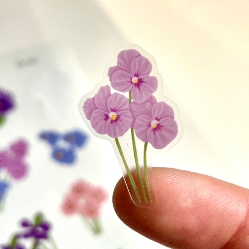 May include: Three purple flowers with yellow centers on clear sticker paper. The flowers are arranged in a bouquet with green stems.
