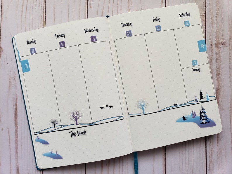 May include: A weekly planner with a winter theme. The planner has a blue cover and is open to the week of December 17th. The days of the week are labeled at the top of each column. The planner has a wintery design with trees, snow, and a snowflake.