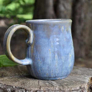 May include: A handmade ceramic mug with a blue and brown glaze. The mug has a rounded body and a large, curved handle. It is sitting on a tree stump outdoors.