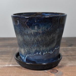 May include: A ceramic planter with a dark blue glaze and a matching saucer. The planter has a flared shape and a glossy finish. The glaze has a textured, streaked appearance with lighter blue and white accents. A decorative item for plants.