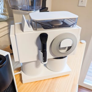 Könnte beinhalten: Weiße Kaffeemaschine mit einem durchsichtigen Deckel und einer schwarzen Bürste, die an der Seite befestigt ist. Die Kaffeemaschine hat einen weißen Sockel und einen silbernen Ring um den oberen Teil. Die Bürste ist mit "Fellow" beschriftet.