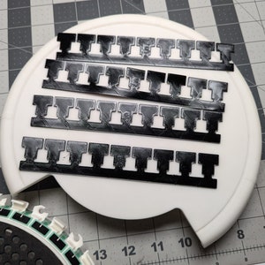 Op de afbeelding: Vier zwarte plastic strips met een herhalend patroon van kleine vierkanten en rechthoeken. De strips zijn gestapeld op een wit oppervlak.