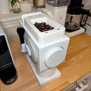 Könnte beinhalten: Weiße elektrische Kaffeemühle mit einem durchsichtigen Behälter, gefüllt mit dunkel gerösteten Kaffeebohnen. Die Mühle hat ein weißes Zifferblatt an der Seite.