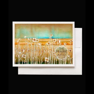 May include: A greeting card featuring a watercolor painting of white wildflowers in a field. The artwork has a warm color palette with orange, yellow, and blue hues. The card is accompanied by a white envelope. The text "THE PRAIRIE ARTIST" is visible.