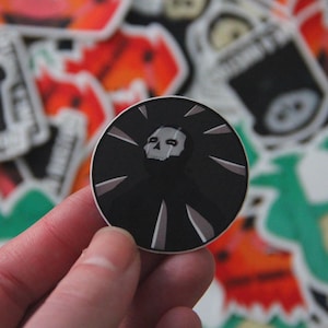 May include: A black and white sticker with a skull and crossbones design. The skull is white and the crossbones are black. The sticker is surrounded by a black background with white lines radiating out from the center.