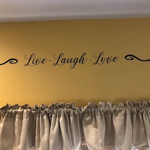 May include: A wall decal with the text "Live. Laugh. Love." in a cursive font. The decal is placed on a yellow wall with a white ceiling. The decal is centered on the wall and is surrounded by a decorative border.
