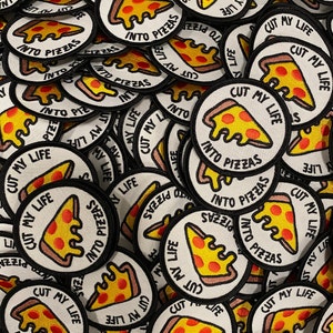 May include: A pile of round, white fabric patches with black outlines. Each patch features a yellow and red pizza slice with the text "Cut My Life Into Pizzas".