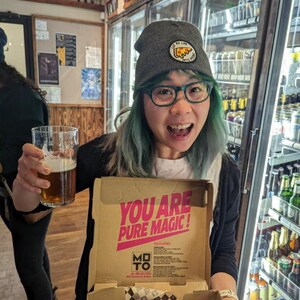 May include: A person wearing a gray beanie with a pizza slice graphic and green hair holds a glass of beer and a pizza box. The box is open and shows a square pizza with cheese and sauce. The box has the text "You are pure magic!" printed on it.