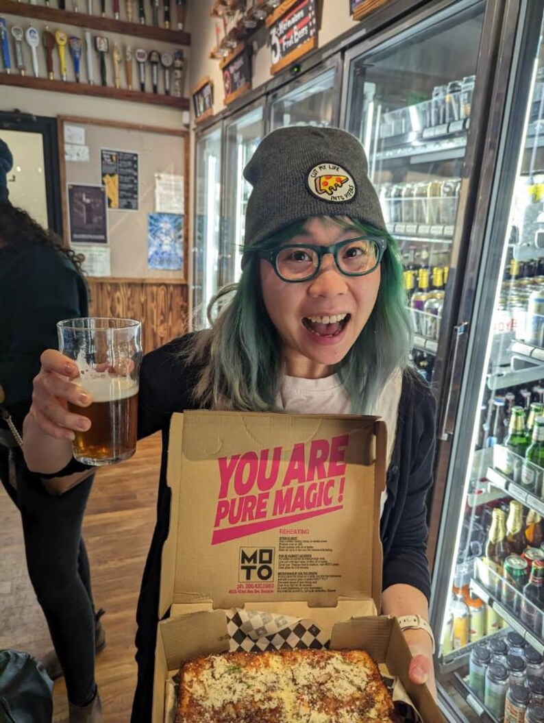 May include: A person wearing a gray beanie with a pizza slice graphic and green hair holds a glass of beer and a pizza box. The box is open and shows a square pizza with cheese and sauce. The box has the text "You are pure magic!" printed on it.