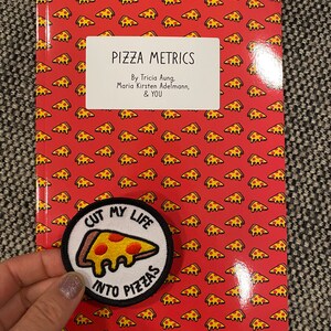 May include: A red book with a pizza pattern on the cover. The book title is "Pizza Metrics" and the authors are Tricia Aung, Maria Kirsten Adelmann, and "You". A white patch with a pizza slice and the text "Cut my life into pizzas" is attached to the front cover.