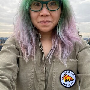 May include: A person with green and purple hair is wearing a green shirt with a patch that says "Cut my life into pizzas".