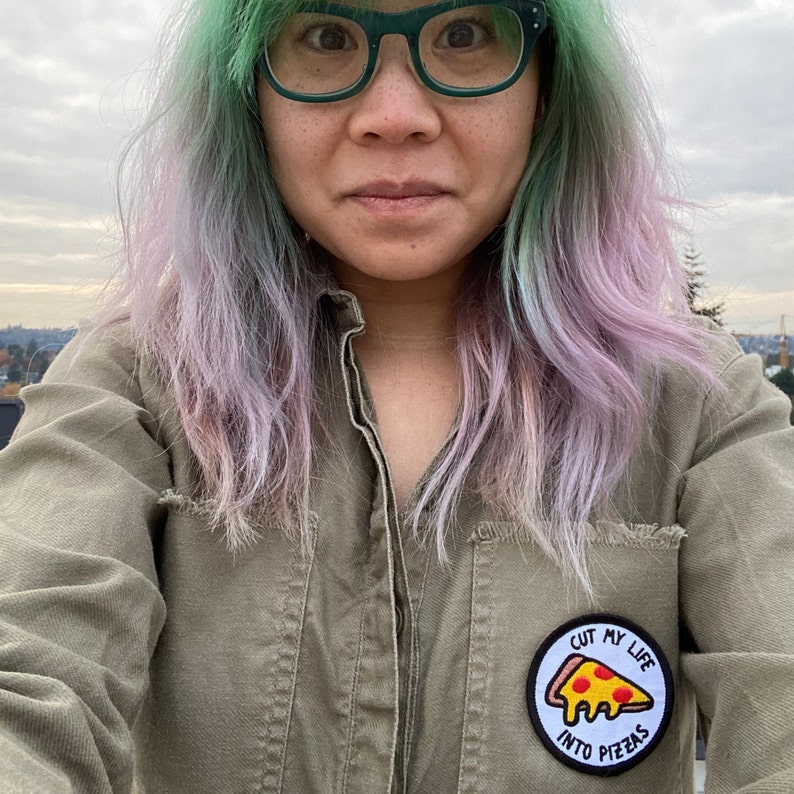 May include: A person with green and purple hair is wearing a green shirt with a patch that says "Cut my life into pizzas".