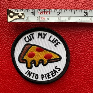 May include: A white round embroidered patch with black stitching. The patch features a pizza slice with red pepperoni and the text "CUT MY LIFE INTO PIZZAS".