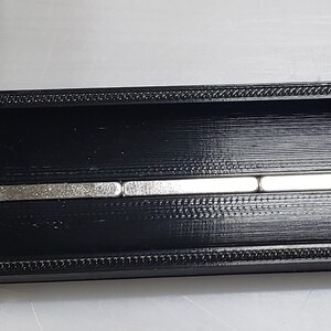 May include: A black plastic track with a series of five silver magnets embedded in the centre. The track is approximately 5 cm long.