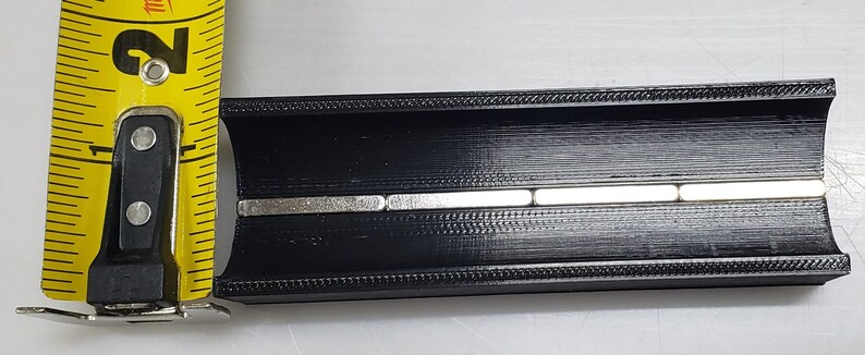 May include: A black plastic track with a series of five silver magnets embedded in the centre. The track is approximately 5 cm long.