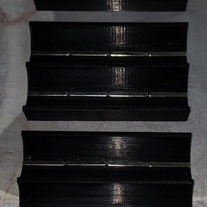 May include: Three black plastic organisers with multiple rows of magnets. The organisers are stacked on top of each other. A yellow tape measure is visible at the top of the image.
