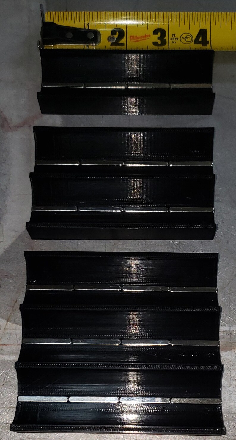 May include: Three black plastic organisers with multiple rows of magnets. The organisers are stacked on top of each other. A yellow tape measure is visible at the top of the image.