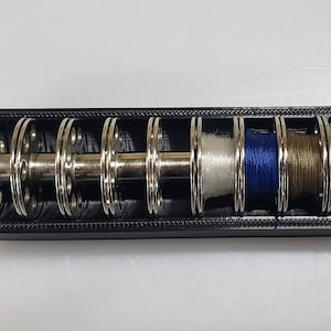 May include: A black plastic spool holder with six metal spools, each holding a different colour thread: white, blue, brown, and green.