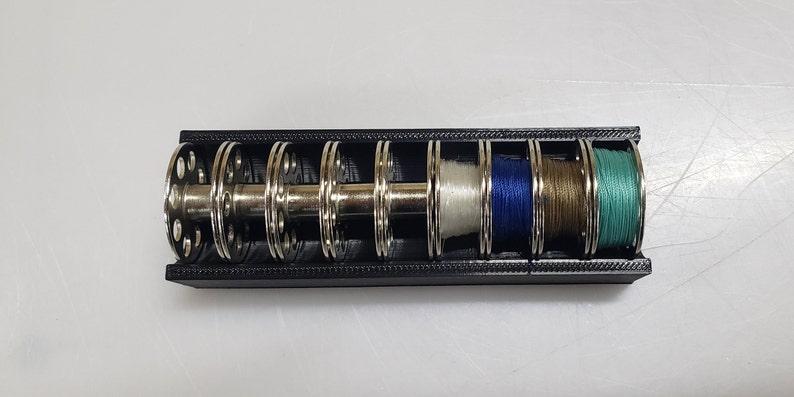 May include: A black plastic spool holder with six metal spools, each holding a different colour thread: white, blue, brown, and green.