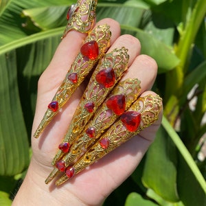 Op de afbeelding: Set van vijf gouden ringen met rode hartvormige strass steentjes. De ringen zijn ontworpen om eruit te zien als lange, puntige nagels.