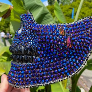 May include: A skull-shaped mask covered in iridescent blue and black rhinestones. The mask is made of a hard material and has a detailed design.