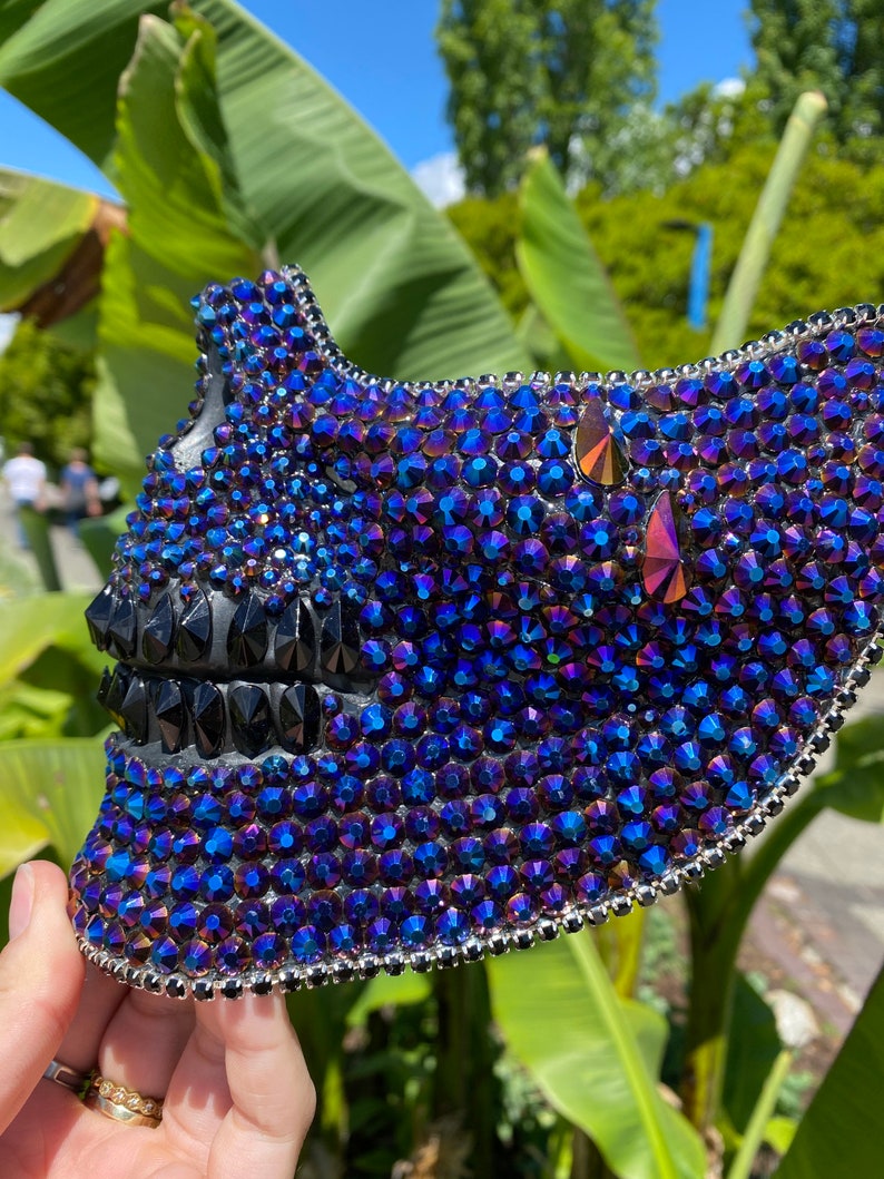 May include: A skull-shaped mask covered in iridescent blue and black rhinestones. The mask is made of a hard material and has a detailed design.