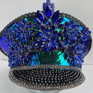 以下が含まれることがあります： 黒い縁と青と緑のスパンコールで覆われた王冠が付いた装飾的な帽子。 帽子は、花柄に配置された大きく輝く青い宝石で飾られており、魅力的で人目を引くデザインを作成しています。