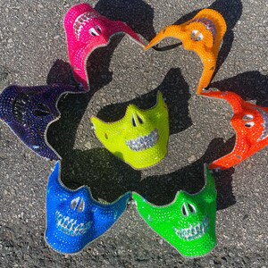 Op de afbeelding: Zeven kleurrijke schedelmaskers met strass. De maskers zijn gemaakt van plastic en hebben verschillende kleuren, waaronder roze, oranje, blauw, groen en paars.