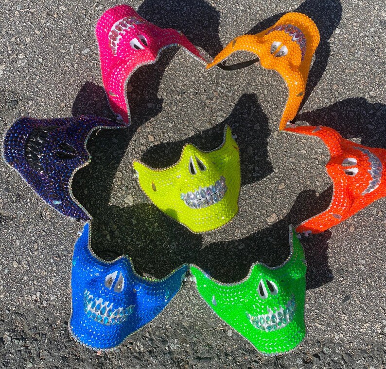 Op de afbeelding: Zeven kleurrijke schedelmaskers met strass. De maskers zijn gemaakt van plastic en hebben verschillende kleuren, waaronder roze, oranje, blauw, groen en paars.