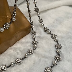 May include: A silver-toned necklace with alternating faux pearls and sparkling floral-shaped embellishments. The necklace is displayed against a soft, white background, showcasing its delicate design and intricate details.
