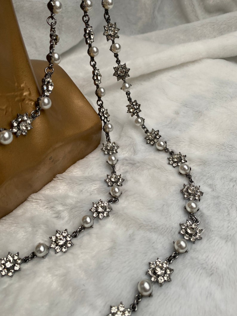 May include: A silver-toned necklace with alternating faux pearls and sparkling floral-shaped embellishments. The necklace is displayed against a soft, white background, showcasing its delicate design and intricate details.