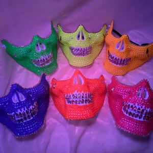 Op de afbeelding: Zes schedelmaskers gemaakt van strass in verschillende kleuren, waaronder groen, geel, oranje, rood en blauw. De maskers hebben een witte basis en zilveren strasssteentjes voor de tanden.