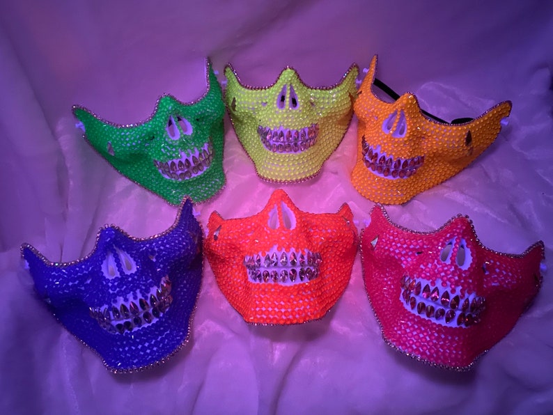Op de afbeelding: Zes schedelmaskers gemaakt van strass in verschillende kleuren, waaronder groen, geel, oranje, rood en blauw. De maskers hebben een witte basis en zilveren strasssteentjes voor de tanden.