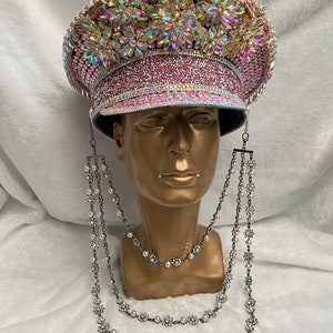 May include: A bedazzled pink and gold military-style hat with iridescent rhinestones and floral accents. The hat is displayed on a gold mannequin head and is adorned with a silver-toned, layered necklace with pearl and rhinestone floral details.