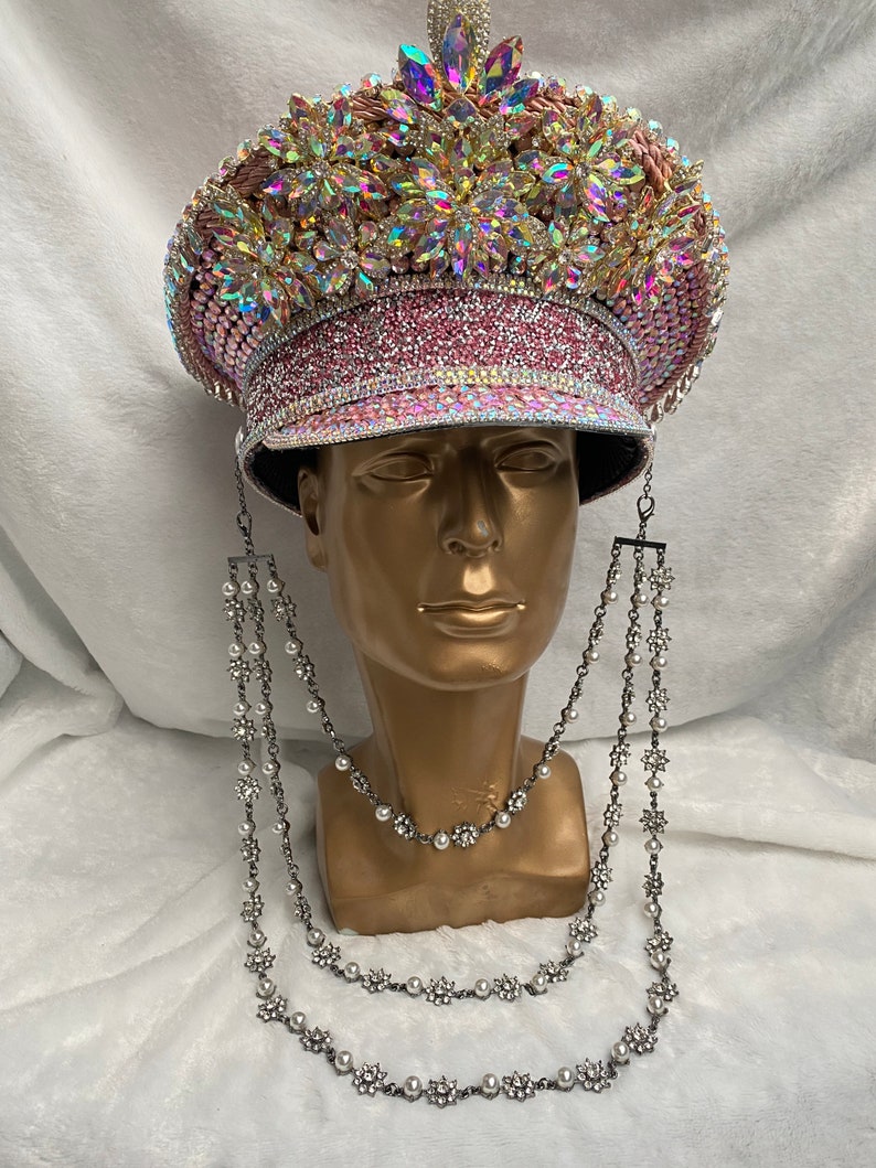 May include: A bedazzled pink and gold military-style hat with iridescent rhinestones and floral accents. The hat is displayed on a gold mannequin head and is adorned with a silver-toned, layered necklace with pearl and rhinestone floral details.