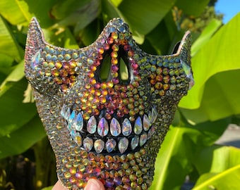 May include: A black skull mask covered in iridescent rhinestones. The rhinestones are in shades of gold, orange, and purple.