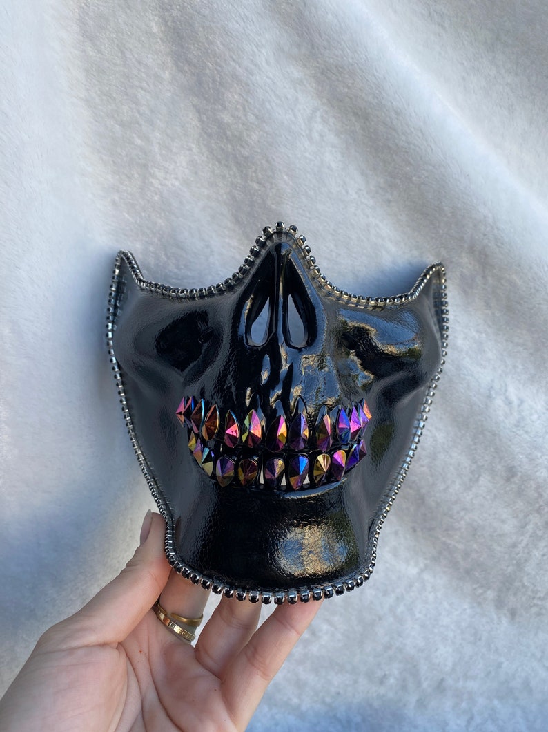 May include: Black skull mask with iridescent crystal teeth and silver beaded trim. The mask has a glossy finish and is designed to cover the lower half of the face. A unique accessory for costume or cosplay.