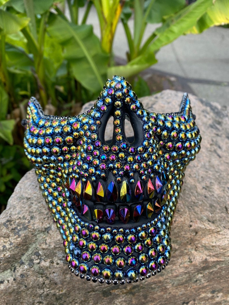 May include: A black mask covered in iridescent, multicolored rhinestones. The mask has a jagged, tooth-like design with colorful gems. The mask is resting on a gray rock, with green foliage in the background.