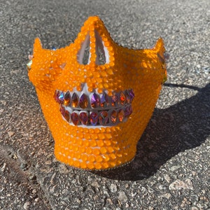 Op de afbeelding: Oranje schedelmasker bedekt met strass met een rij regenboogkleurige stenen als tanden.